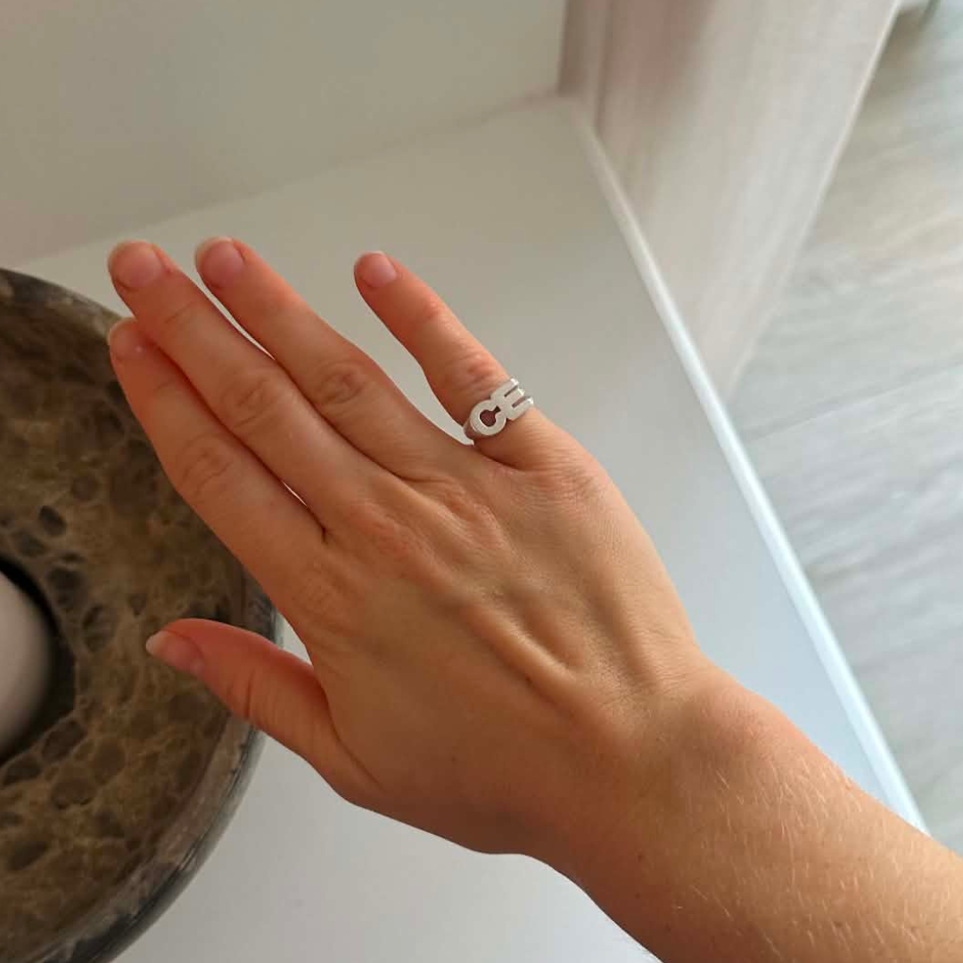 A close-up of a person's hand wearing a silver ring with yhe lertter C E design, set against a minimalist indoor background.