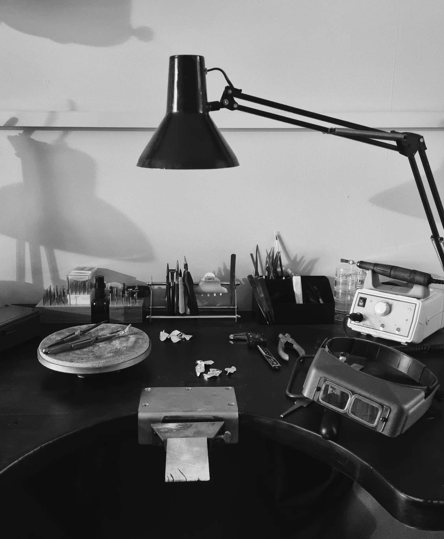 Black and white photo of a desk with various items including a lamp, tools, and electronic equipment.